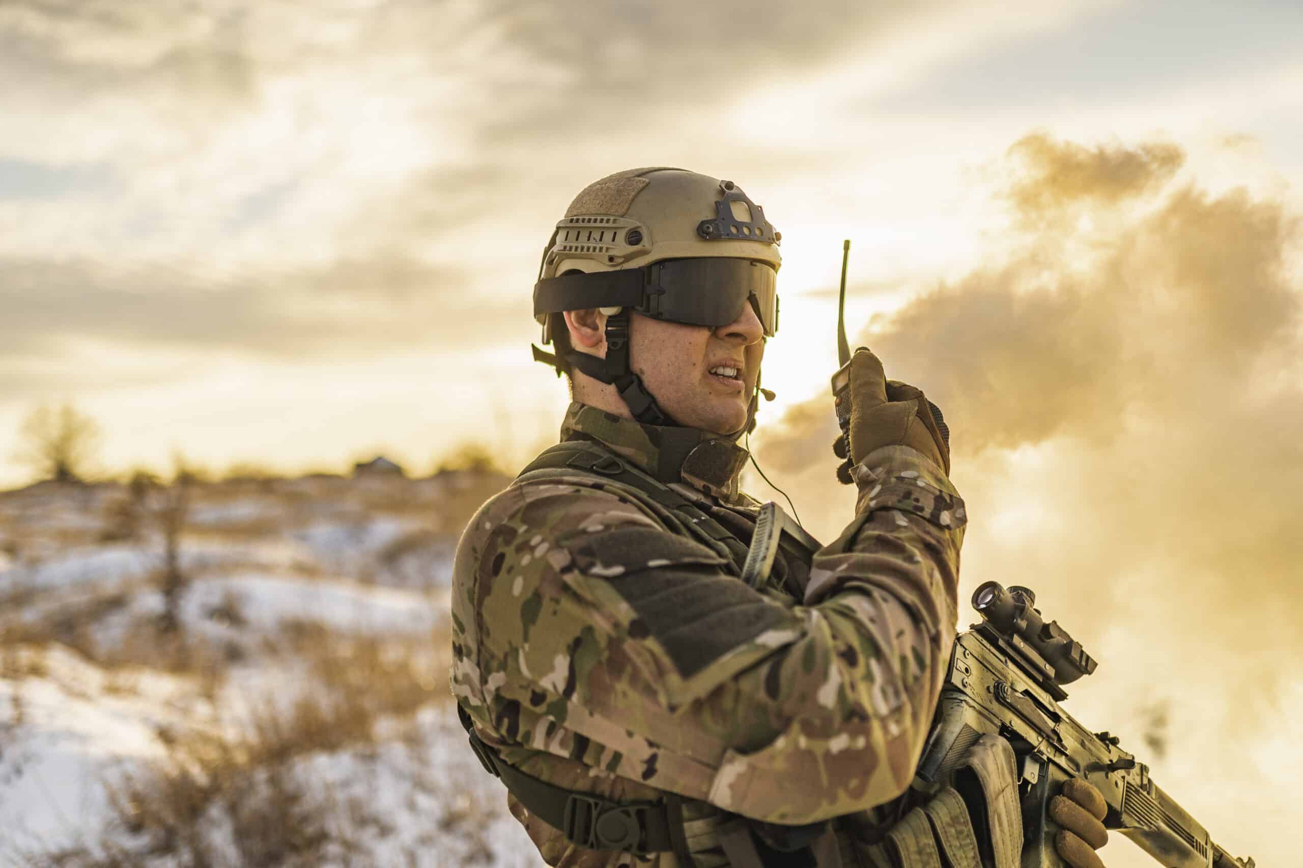 Military soldier in full tactical gear holding a rifle, wearing camouflage uniform, helmet, and goggles during outdoor training exercise.