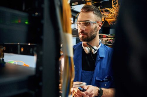 A male technician wearing safety glasses and headphones working on server equipment in a high-tech data center. Focused on network and infrastructure maintenance.